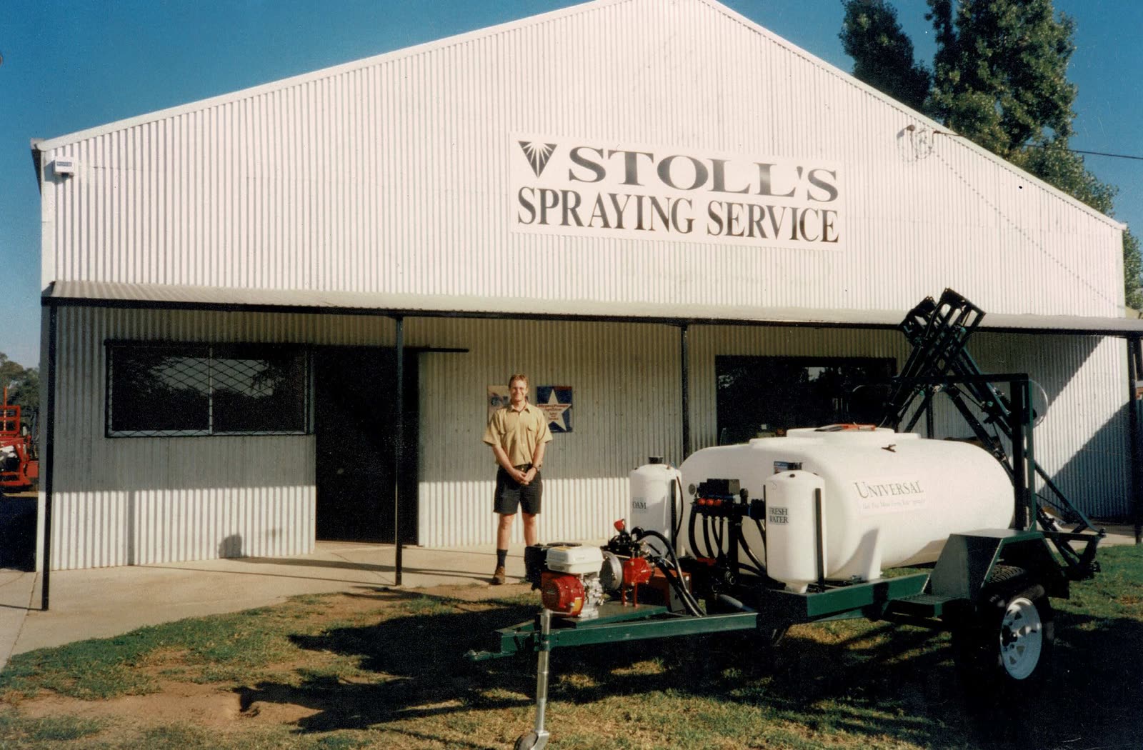 A man standing beside a small trailed spray unit outside a shed signed Stoll’s Spraying Service